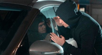 Persona con capucha mirando al interior de un coche por la ventanilla de noche, ilustrando el riesgo de robo de vehículo o de piezas de carrocería en zonas urbanas.